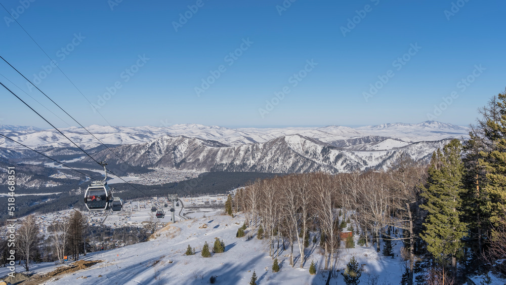 Funicular over a snowy valley. The booths are visible on the ropes. The forest is below. A picturesque mountain range against a clear blue sky. Altai. Manzherok