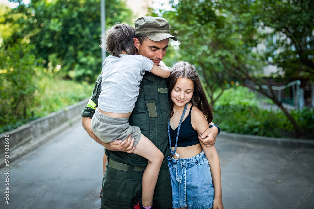 Military man in olive uniform and cap hugging his two kids near house outdoors in summer