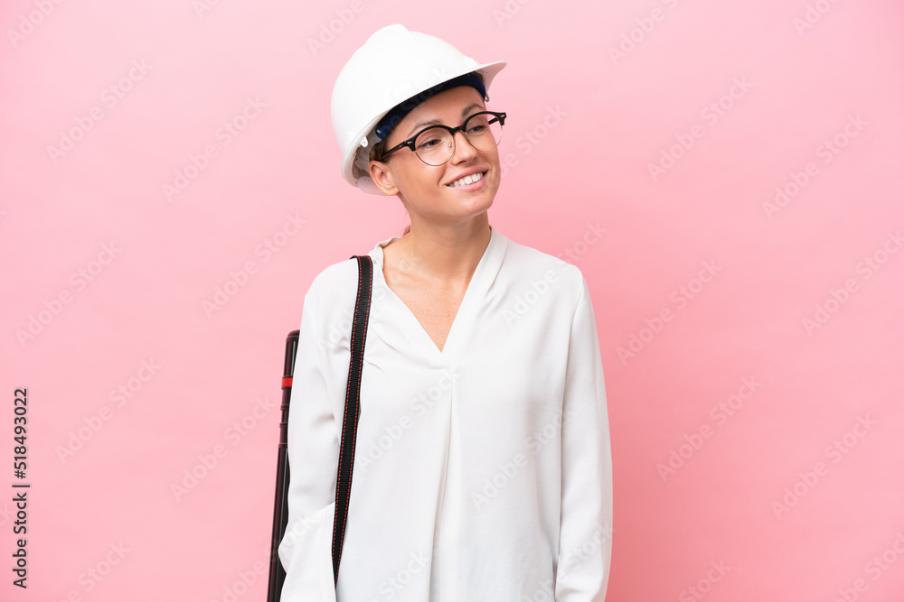 Young architect Russian woman with helmet and holding blueprints isolated on pink background looking to the side and smiling