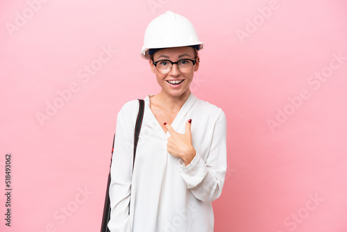Young architect Russian woman with helmet and holding blueprints isolated on pink background with surprise facial expression