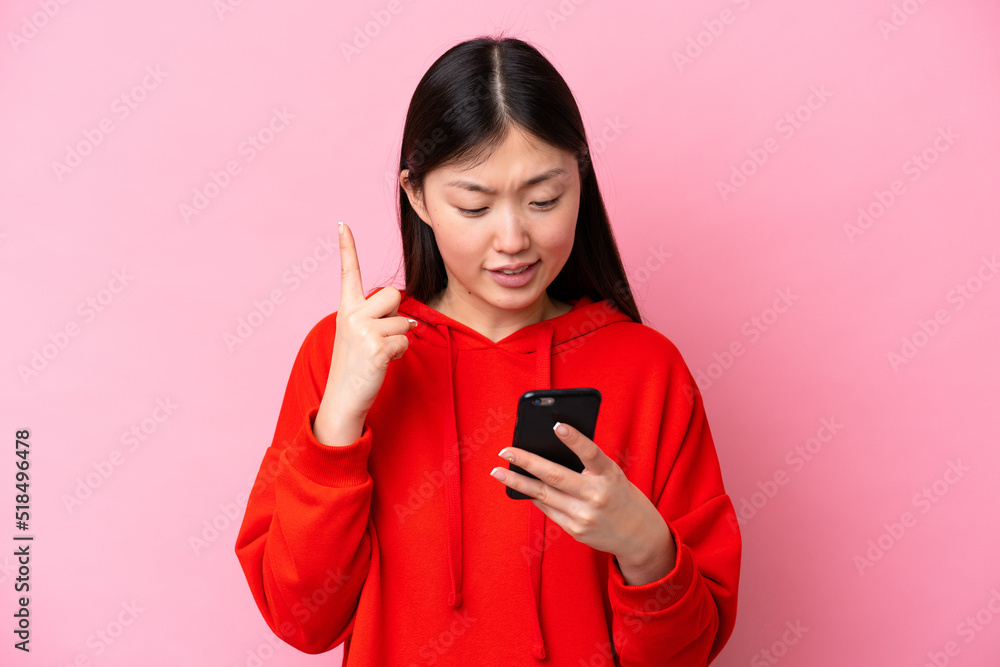 Young Chinese woman isolated on pink background using mobile phone and lifting finger Young Chinese woman isolated on pink background using mobile phone and lifting finger