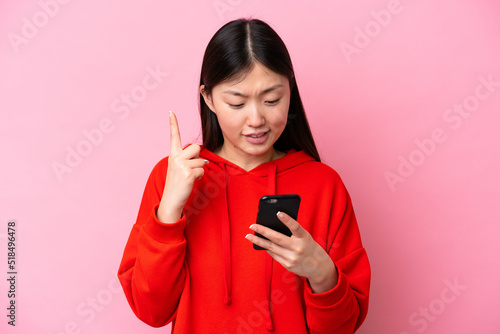 Young Chinese woman isolated on pink background using mobile phone and lifting finger