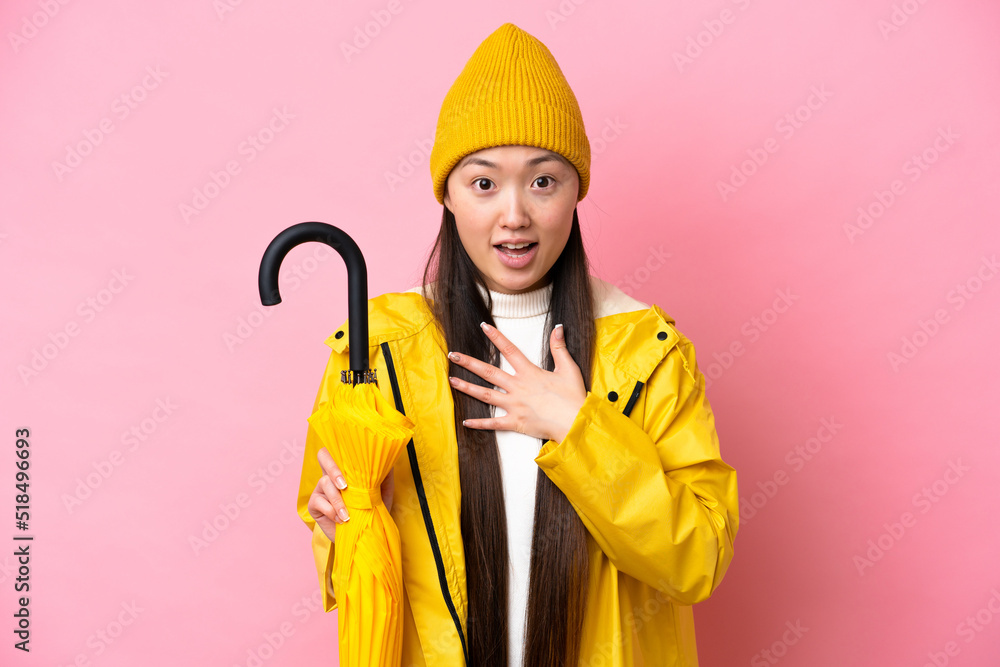 Young Chinese woman with rainproof coat and umbrella isolated on pink background surprised and shocked while looking right Young Chinese woman with rainproof coat and umbrella isolated on pink background surprised and shocked while looking right