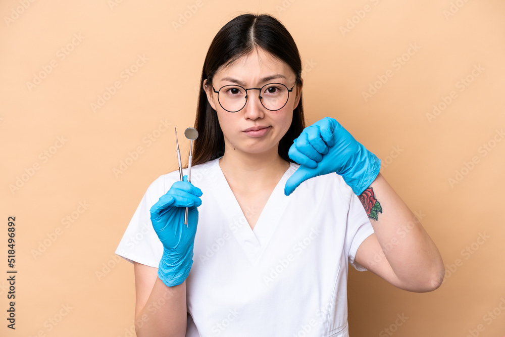 Dentist Chinese woman holding tools isolated on beige background showing thumb down with negative expression Dentist Chinese woman holding tools isolated on beige background showing thumb down with negative expression