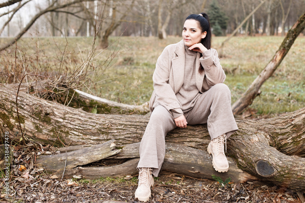 Fototapeta premium portrait of brunette woman in beige coat with beautiful long hair sitting on a tree in the park