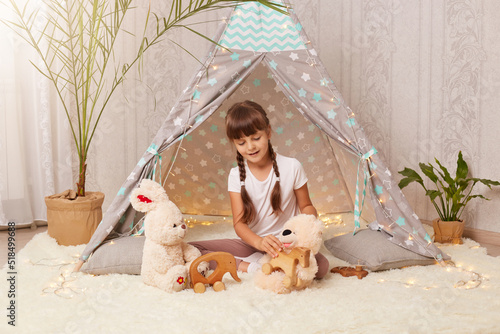 Indoor shot of charming dark haired Caucasian little girl wearing white t shirt sitting on floor in wigwam and playing with teddy bear, soft rabbit and eco wooden toys. photo