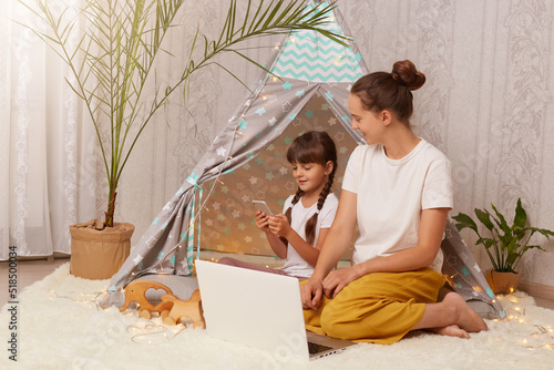 Portrait of dark haired female and her daughter wearing white t shirts sitting in wigwam in front of laptop, child using smart phone, mother looking at kid, family spending time together.