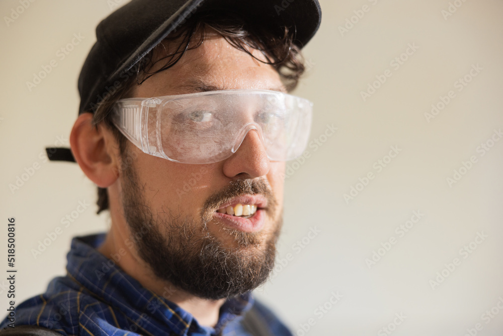 Portrait of a bearded man in overalls and baseball cap at work. Confident contractor standing with arms crossed on a construction object wearing safety glasses. Portrait of a bearded man in overalls and baseball cap at work. Confident contractor standing with arms crossed on a construction object wearing safety glasses.