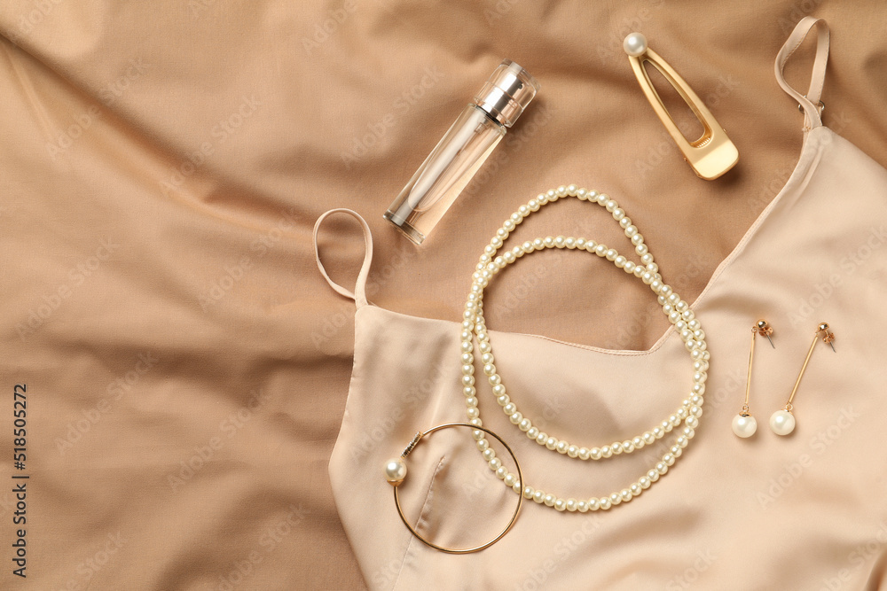 Stylish jewelry with pearls, luxury perfume and silk dress on beige fabric, flat lay Stylish jewelry with pearls, luxury perfume and silk dress on beige fabric, flat lay