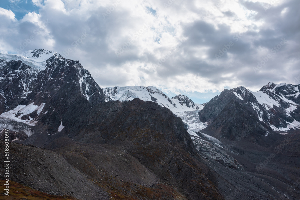 Atmospheric landscape with large snow mountain range with glacier and icefall in dramatic cloudy sky. Awesome high snowy mountains under rainy clouds. White snow on black rocks in overcast weather.