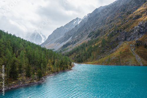 Shiny ripples on turquoise alpine lake against snowy mountain range during rain in changeable weather. Big azure mountain lake in forest valley in vivid autumn colors and snow mountains in overcast.