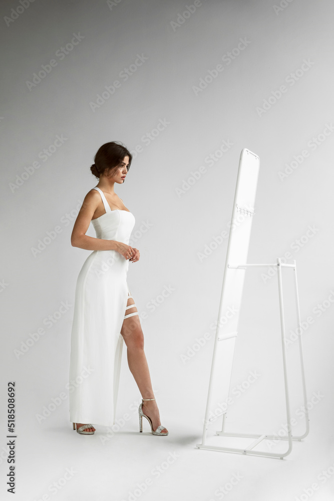 Full length view of young woman with hairstyle in white long dress posing sensual in studio, looking at mirror, isolated grey background. Full length view of young woman with hairstyle in white long dress posing sensual in studio, looking at mirror, isolated grey background.