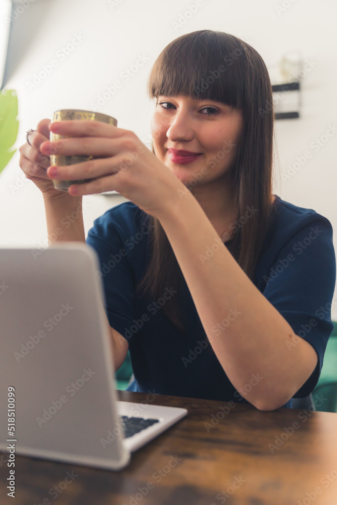 Smiling white woman with dark hair bangs and soft makeup sitting by a desk with a laptop holding a mug looking at the camera. Indoor vertical shot. High quality photo Smiling white woman with dark hair bangs and soft makeup sitting by a desk with a laptop holding a mug looking at the camera. Indoor vertical shot. High quality photo