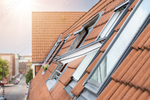 Open ventilation waterproof rooftop window exterior against sunny sky light. Velux style roof with red brick tiles. European city street attic mansard modern roofwindow service, install maintenance photo