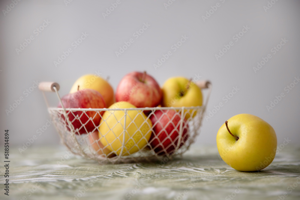 Pile of yellow and red apples lying on the table in a wire basket with white background and one apple lies nearby. Yellow and red apples in a basket on a table Pile of yellow and red apples lying on the table in a wire basket with white background and one apple lies nearby. Yellow and red apples in a basket on a table