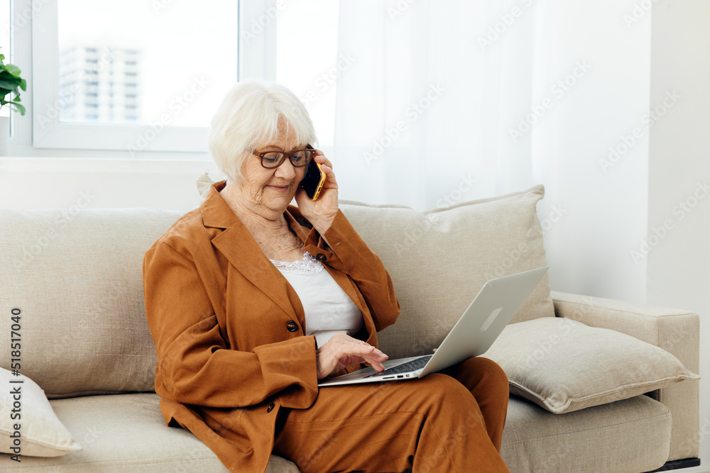 a happy pleasant elderly woman is sitting in a comfortable environment on a beige sofa in a stylish brown suit working from home with a laptop holding it on her lap a happy pleasant elderly woman is sitting in a comfortable environment on a beige sofa in a stylish brown suit working from home with a laptop holding it on her lap