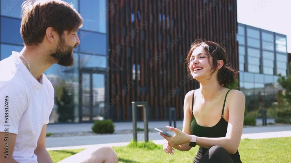 Cheerful happy Caucasian couple giving high five to each other and laughing while sitting on green grass on summer sunny day. Finishing workout. Man and woman end doing sport exercising. Cheerful happy Caucasian couple giving high five to each other and laughing while sitting on green grass on summer sunny day. Finishing workout. Man and woman end doing sport exercising.