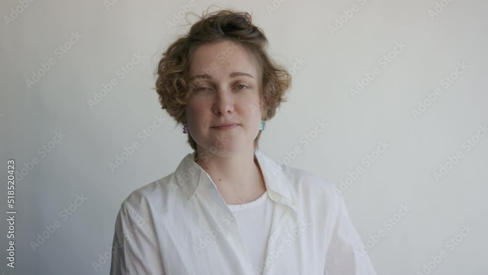 Young cheerful happy smiling woman with short curly hair take professional photo portrait in studio. Female in casual shirt, sit, bite lips, shy, look at camera on white background in modern photo lab Young cheerful happy smiling woman with short curly hair take professional photo portrait in studio. Female in casual shirt, sit, bite lips, shy, look at camera on white background in modern photo lab