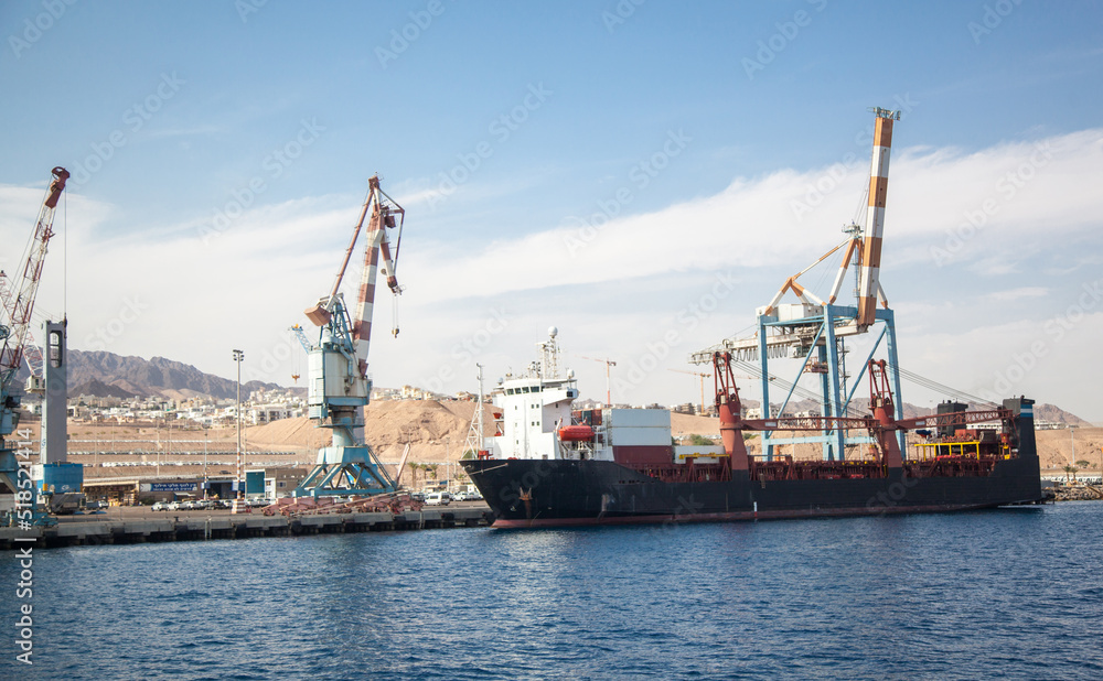 A huge cargo ship is anchored in a cargo port. The cranes are ready to be loaded on board the cargo ship for dispatch. International trade and logistics. A huge cargo ship is anchored in a cargo port. The cranes are ready to be loaded on board the cargo ship for dispatch. International trade and logistics.
