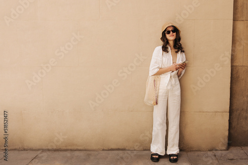 Full length cute caucasian woman holding smartphone stands near city wall with space for text. Brunette with wavy hair wears casual clothes. Cell phone usage concept