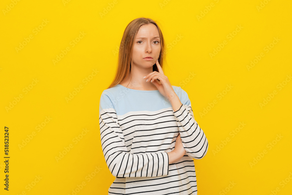 Portrait of thoughtful female, keeps finger on cheek, contemplates about future wedding with boyfriend or making important decision, dressed in casual striped sweatshirt, yellow background Portrait of thoughtful female, keeps finger on cheek, contemplates about future wedding with boyfriend or making important decision, dressed in casual striped sweatshirt, yellow background