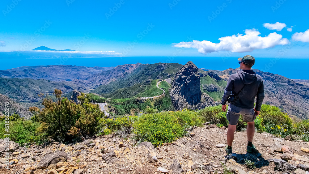 Man with scenic view on mountain road next to volcanic rock formation Roque de Agando in Garajonay National Park on La Gomera, Canary Islands, Spain, Europe. Seen from lookout Mirador Morro de Agando Man with scenic view on mountain road next to volcanic rock formation Roque de Agando in Garajonay National Park on La Gomera, Canary Islands, Spain, Europe. Seen from lookout Mirador Morro de Agando