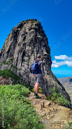 Sport man with backpack standing in front of massive volcanic rock formation Roque de Agando in Garajonay National Park on La Gomera, Canary Islands, Spain, Europe. Hiking trail on sunny day in summer