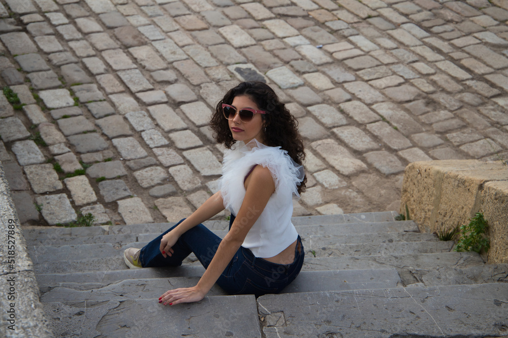 beautiful young woman with dark, curly hair sitting on a staircase in a park. The woman wears sunglasses and makes different expressions. Expressions and lifestyle concept beautiful young woman with dark, curly hair sitting on a staircase in a park. The woman wears sunglasses and makes different expressions. Expressions and lifestyle concept