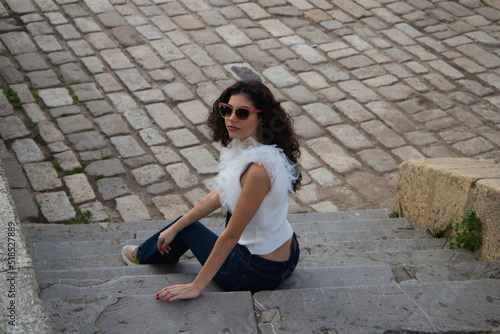 beautiful young woman with dark, curly hair sitting on a staircase in a park. The woman wears sunglasses and makes different expressions. Expressions and lifestyle concept © @skuder_photographer