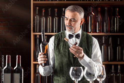 Sommelier with a glass of wine. Examination of wine products. Restaurant staff, expert wine steward among shelves of wine bottles. Stylish middle-aged man with a grey beard photo