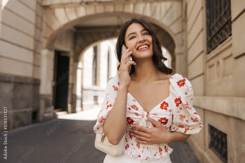 Happy young caucasian woman answering phone standing on street. Brunette with wavy hair wears white blouse. Emotion concept, use Happy young caucasian woman answering phone standing on street. Brunette with wavy hair wears white blouse. Emotion concept, use