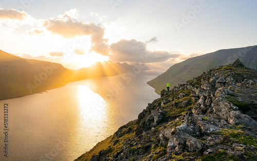 Crazy hiker in yellow jacket umping between rocks. Marvelous colorful daybreak in rocky mountains, heavy orange mist in deep valley. Miracle of nature. Klakkur peak, Faroe Islands, Denmark, Europe.