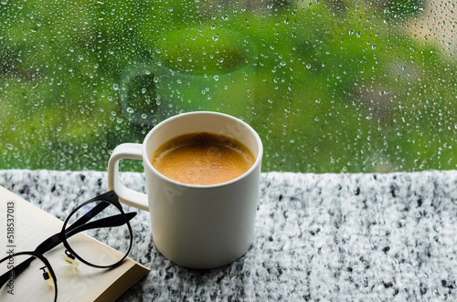 A cup of hot coffee with book and spectacles on table in morning with rain drop on window and green background. Stay home and relaxing concept.
