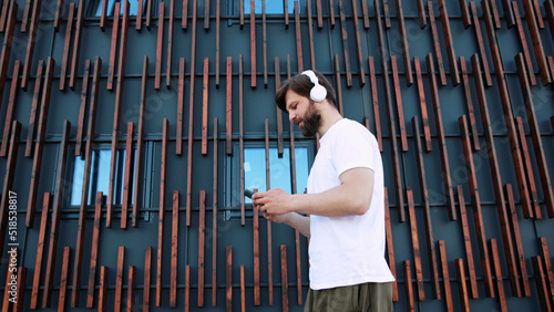 High quality photo of handsome Caucasian man in headphones walking the street, listening to music and texting mesage on smartphone. Male messaging online on mobile phone while stepping. Outdoors. photo