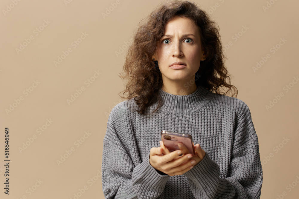 Confused unhappy curly lady in gray casual sweater read conflicting information in message posing isolated on over beige background. Social media, network, distance communication concept. Copy space Confused unhappy curly lady in gray casual sweater read conflicting information in message posing isolated on over beige background. Social media, network, distance communication concept. Copy space