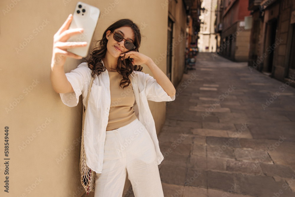 Attractive young blogger girl is broadcasting with smartphone walking in tourist city. Brunette woman with wavy hair wears casual clothes. Social media addiction concept