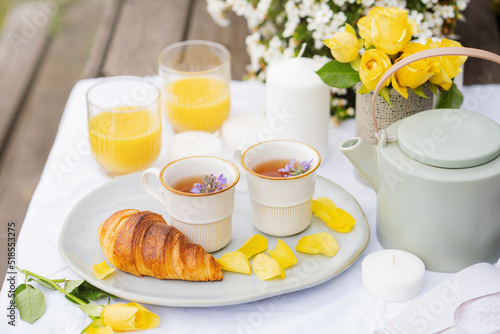 tea party in the garden, herbal tea, teapot, orange juice, croissant, yellow roses and white candles on a white tablecloth