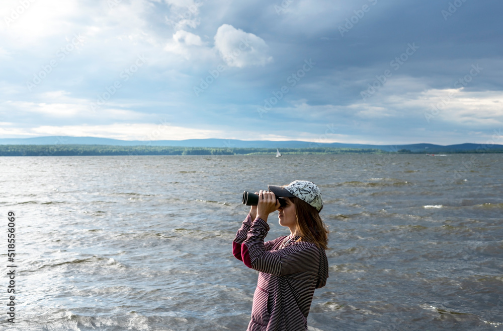 Young woman in cap looking through binoculars at birds on the lake or river Birdwatching, zoology, ecology Research in nature, observation of animals Ornithology copy space