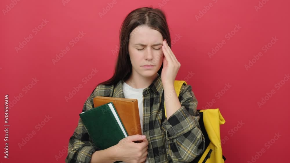 Tired student girl is frowning and touching temple with fingers, has headache or migraine, exhausted of studies, holding books, wearing shirt and yellow backpack, isolated on red background in studio Tired student girl is frowning and touching temple with fingers, has headache or migraine, exhausted of studies, holding books, wearing shirt and yellow backpack, isolated on red background in studio