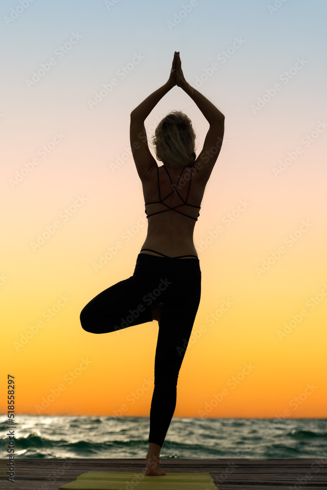 A young woman practices yoga on the ocean coast during sunrise.