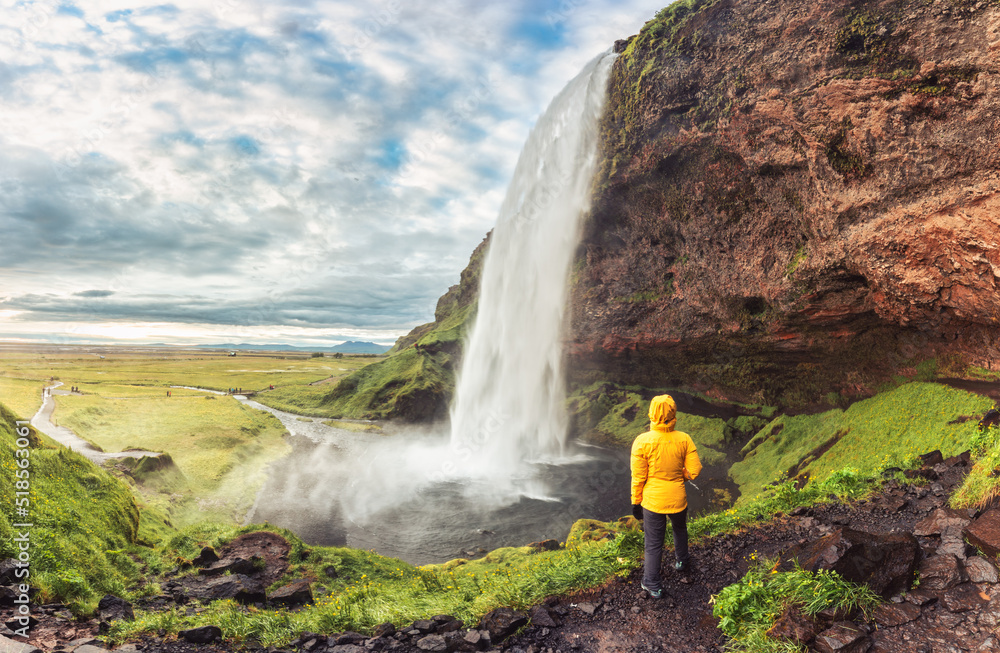 Woman in yellow jacket standing and looking at Seljalandsfoss waterfall flowing from cliff on daylight in summer at South of Iceland