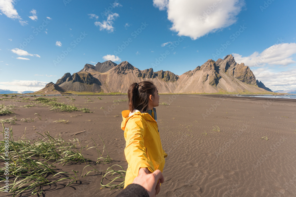 Young asian woman in yellow jacket holding hands with boyfriend on the beach with Vestrahorn mountain in viking village on Stokknes at Iceland