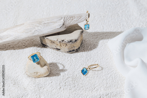 Elegant jewellery set of gold earrings and ring with blue topaz on white background with stones and wood. Minimal style composition. Product still life concept photo