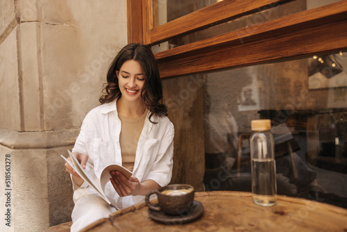 Smiling young caucasian woman leafing through notebook with notes sitting outdoors in cafe. Brunette wears white casual clothes. Student concept