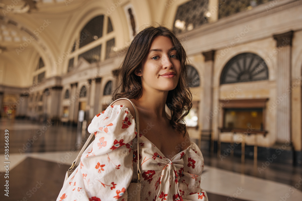 Fototapeta premium Beautiful young caucasian girl looks into camera spending time in large room. Brunette with wavy hair wears blouse summer. City life concept