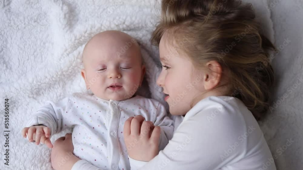 Happy kids, toddler and older sister hugging at home on white blanket, smiling, shot from above. older sister hugs and kisses her newborn sister. Happy kids, toddler and older sister hugging at home on white blanket, smiling, shot from above. older sister hugs and kisses her newborn sister.