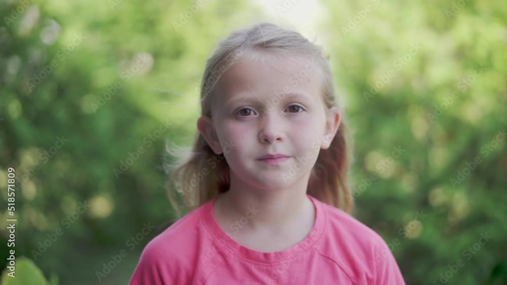 A young girl with blonde stares at the camera in slow motion with green trees as a background. A young girl with blonde stares at the camera in slow motion with green trees as a background.