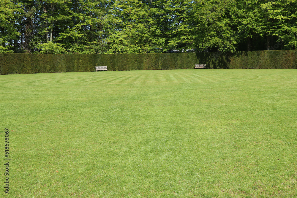 Two park benches by a hedge in a large circular grassy field. Two park benches by a hedge in a large circular grassy field.