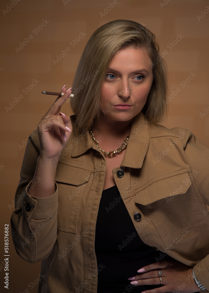 Obraz premium Portrait of a young, beautiful girl with blue eyes and blonde hair in a beige jacket. She looks sternly and smokes a cigarette. She is wearing an expensive watch, a gold chain and rings.