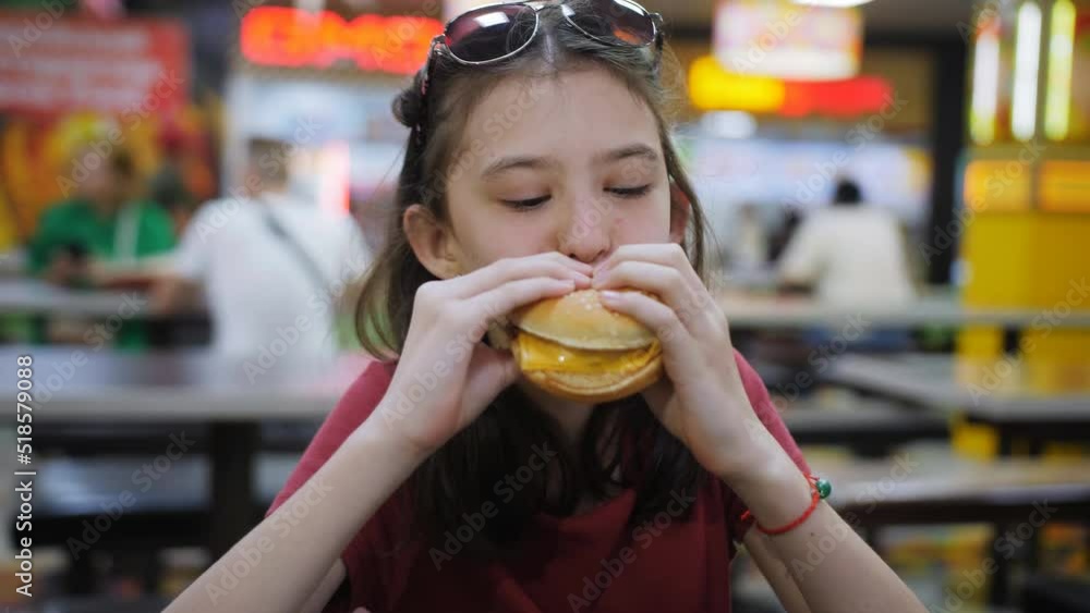 A girl bites a juicy cheeseburger sitting at a table in a diner. A girl bites a juicy cheeseburger sitting at a table in a diner.
